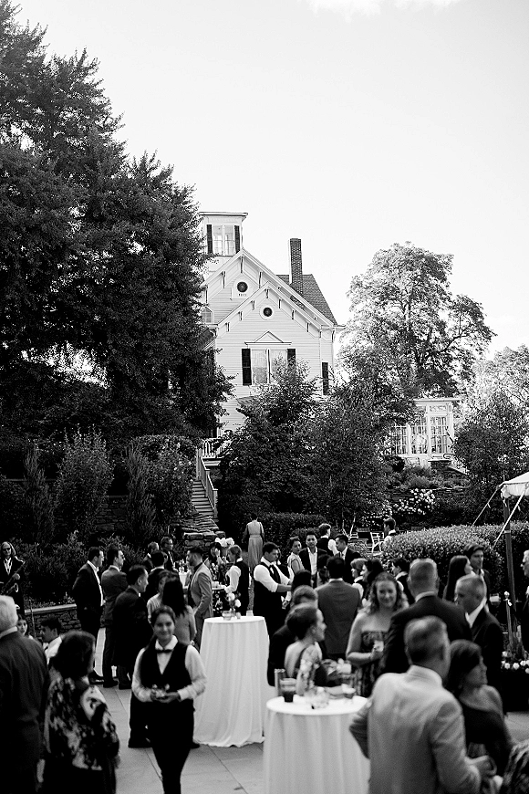 Wedding cocktail hour with guests mingling around white linen cocktail tables, holding drinks on a garden patio near a historic house and tent