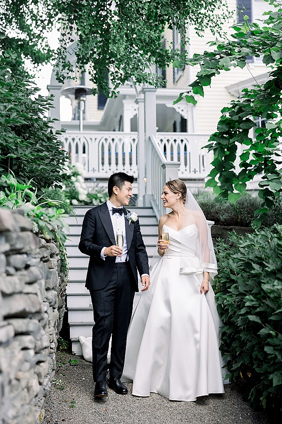 Couple portrait of bride and groom walking on porch steps, holding champagne flutes; she wears a veil and he a tux, by greenery and stone wall