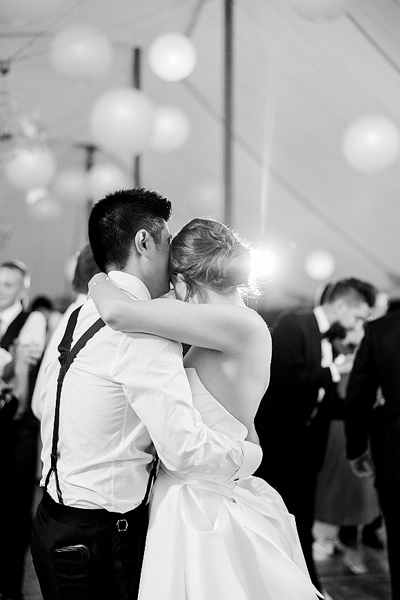 First dance as the newlyweds sway, bride in a strapless wedding dress with updo, groom in suspenders under lantern-lit tent with guests behind