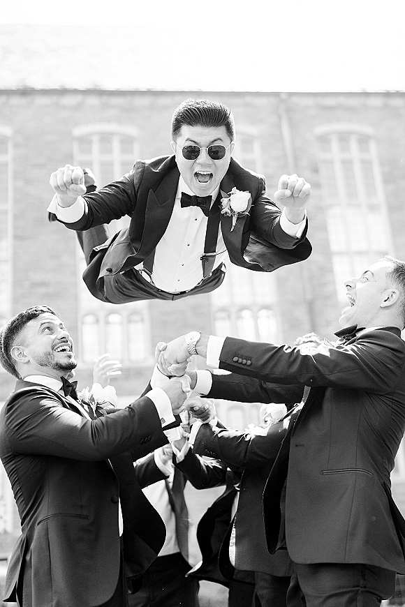 Groomsmen photo of tuxedoed friends tossing the groom into the air, all wearing sunglasses, in a courtyard by a brick building