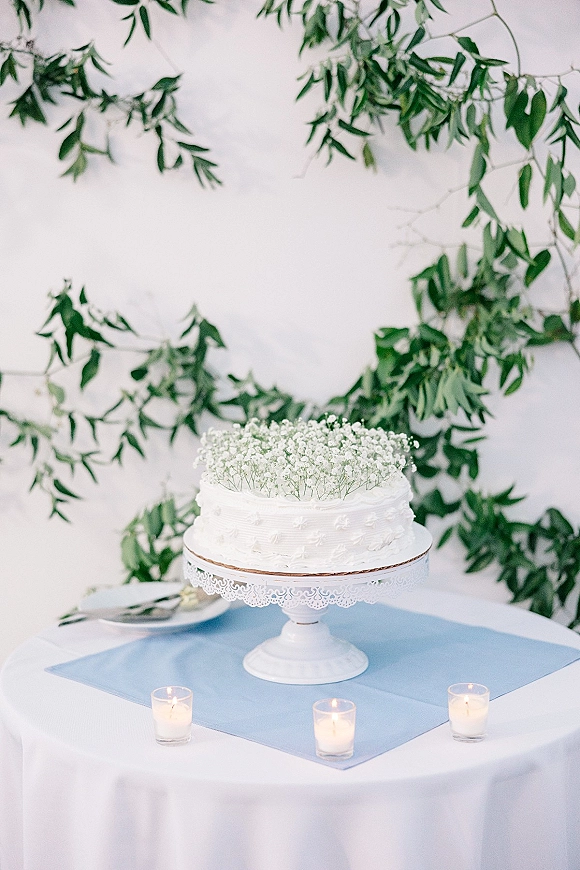 Wedding cake with white buttercream frosting topped with baby's breath on a lace-trimmed stand, with candles and hanging greenery backdrop