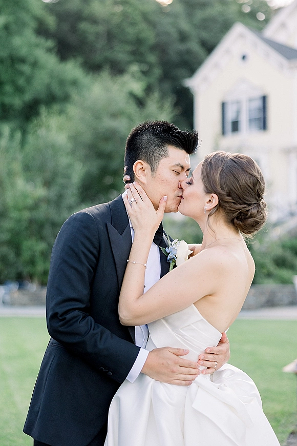 Wedding kiss portrait of bride and groom kissing, bride holding his face as he holds her waist, on green lawn by stone wall and white house