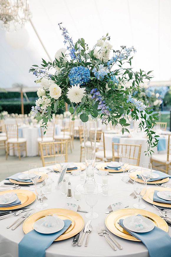 Reception tablescape with wedding table centerpiece in a clear glass vase, blue napkins and gold chargers under chandeliers in a white tent