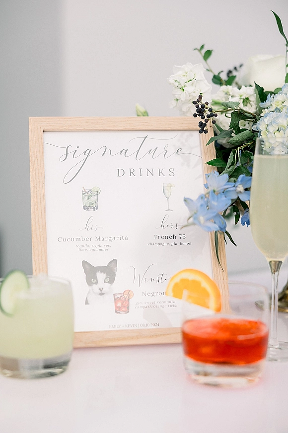 Signature drinks sign in a wood frame with watercolor cocktail menu, blue and white florals, and garnished drinks on a white tabletop