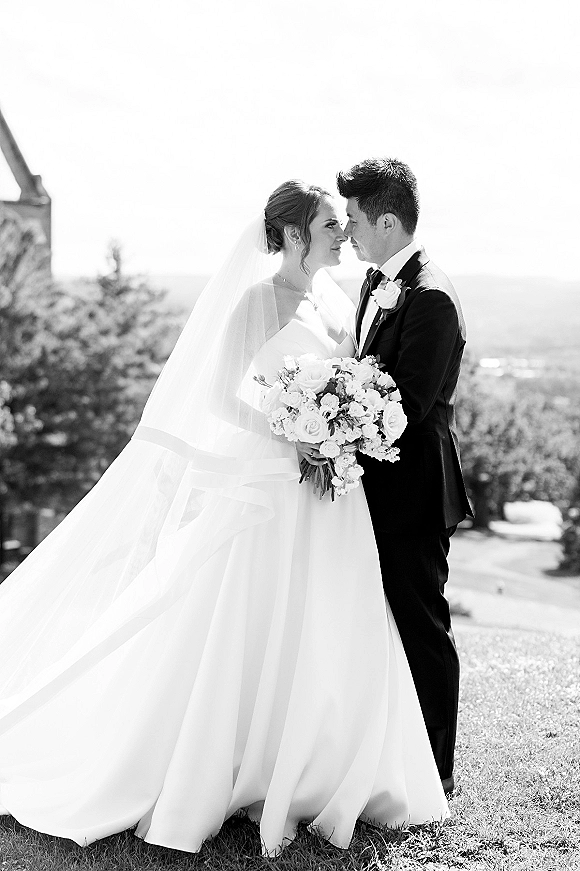Wedding couple portrait in black and white, bride and groom nose to nose with bouquet, her veil blowing on a bright lawn with trees