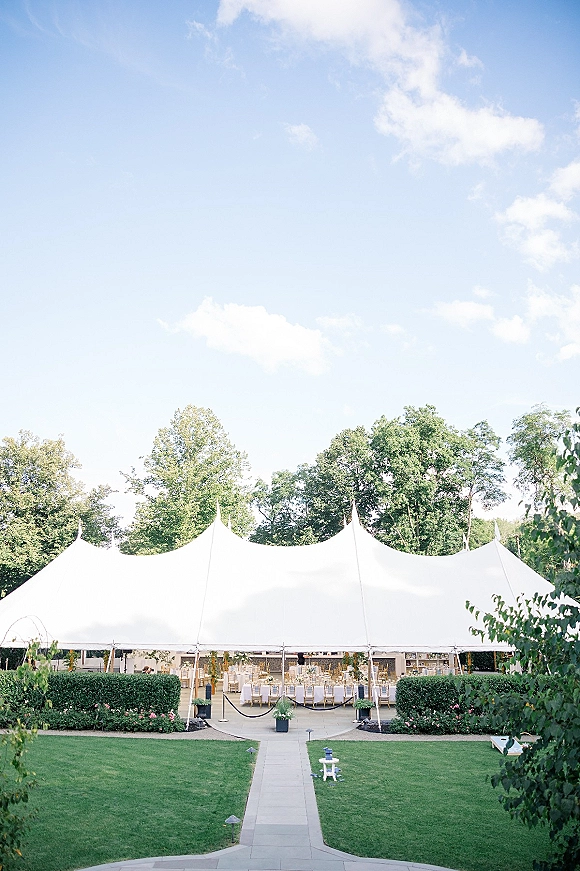 Wedding reception tent with round tables and gold chairs under a white canopy, lined with rope stanchions and pathway lights on a green lawn