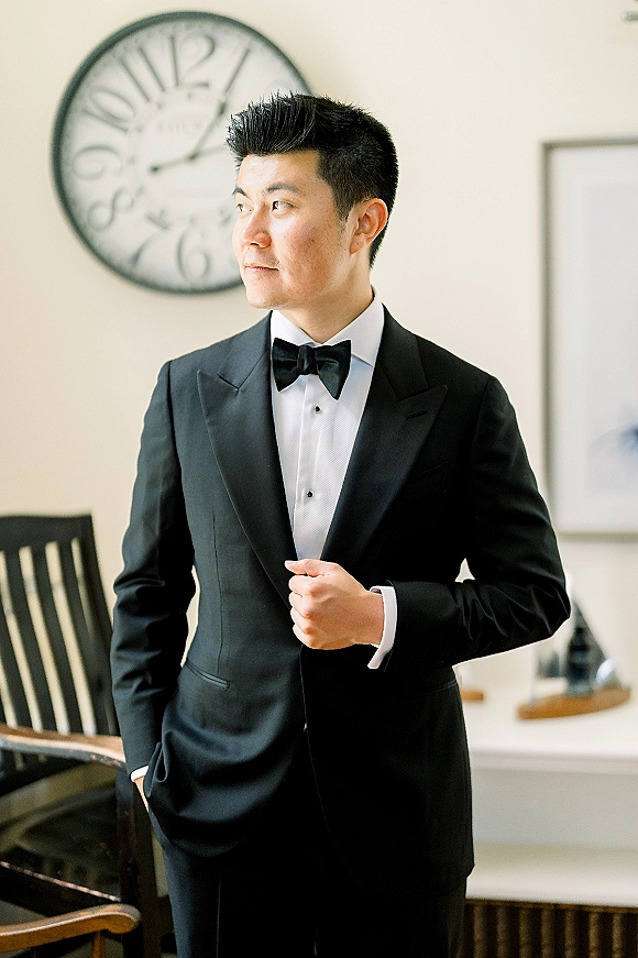 Groom portrait in a black tuxedo with bow tie and pocket square, adjusting his jacket in an indoor room with wall clock and artwork