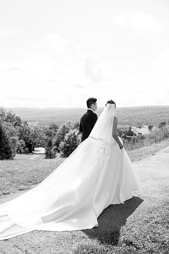 Couple portrait in a black and white wedding photo of bride and groom from behind, walking a path as her long veil trails over hills and valley