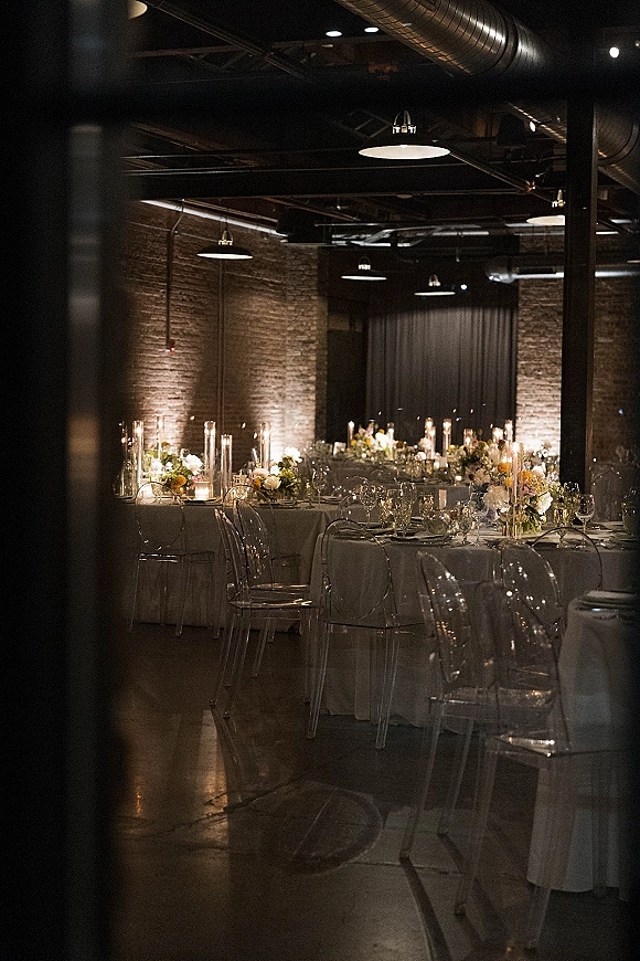 Reception tablescape at an industrial wedding reception with white linens, floral centerpieces, pillar candles, and clear acrylic chairs by brick walls