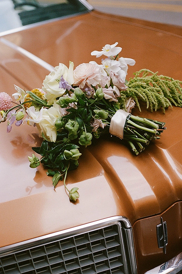 Bridal bouquet resting on a vintage car hood, a wedding bouquet on car with mixed pastel flowers, greenery, and white ribbon wrap