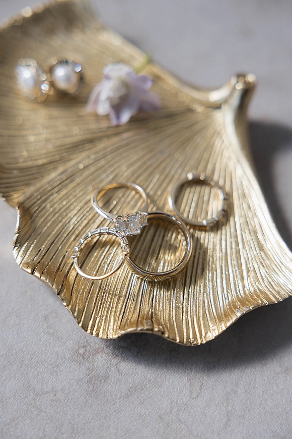 Wedding rings with engagement ring close up on a gold leaf dish beside pearl stud earrings and a tiny flower on light stone surface