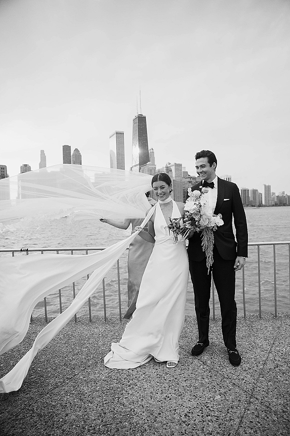 Couple portrait in a black and white wedding portrait style, bride holds bouquet as veil billows by a waterfront skyline railing