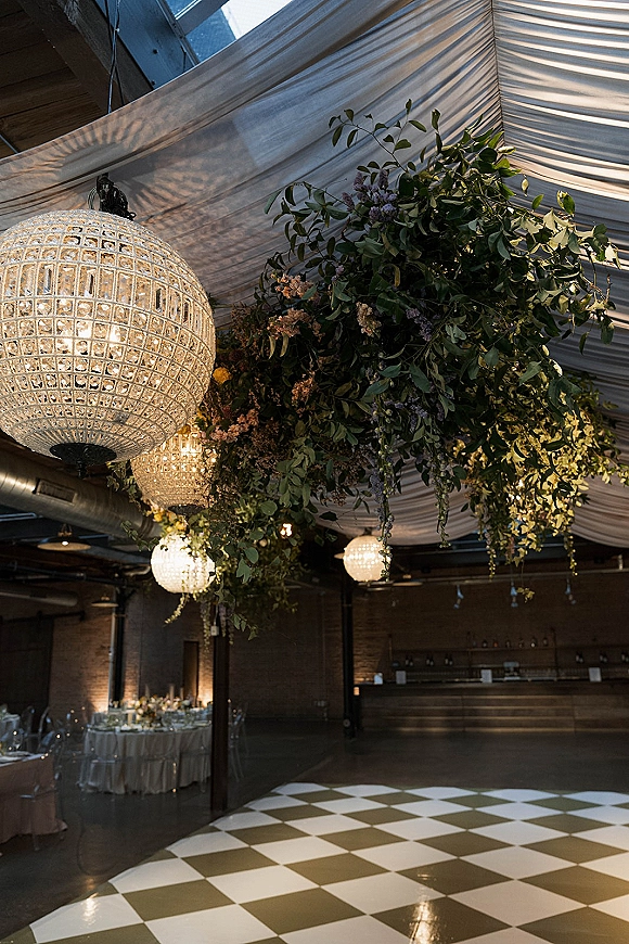 Reception ceiling decor with wedding ceiling draping, hanging greenery, and chandeliers above an industrial brick reception and dance floor