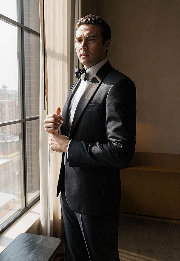 Groom portrait in a black tie tuxedo, wearing a bow tie by a window with sheer curtains and a city skyline beyond