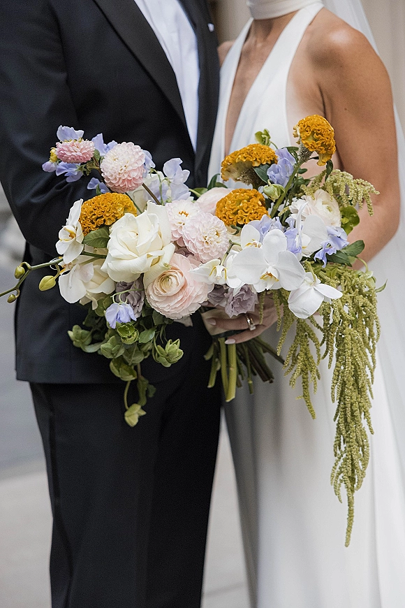 Bridal bouquet with white orchid wedding bouquet blooms, blush and mustard flowers, and trailing amaranthus held by bride and groom indoors