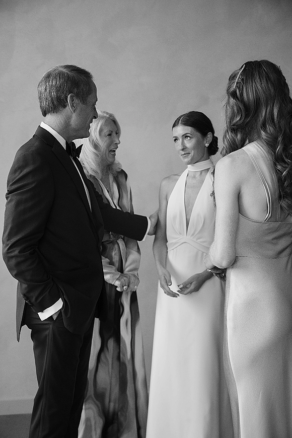 Getting ready moment as bride talks with parents, wearing pearl earrings and hair clip, beside tuxedo and bow tie in plain room