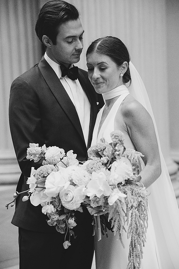 Couple portrait in a black and white wedding portrait style, bride in halter dress with veil and bouquet beside groom in tuxedo by stone columns