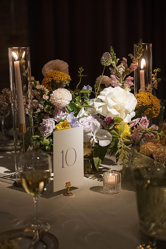 Wedding centerpiece with colorful wedding centerpiece blooms, roses and orchids beside candles in glass hurricane holders on a dark backdrop
