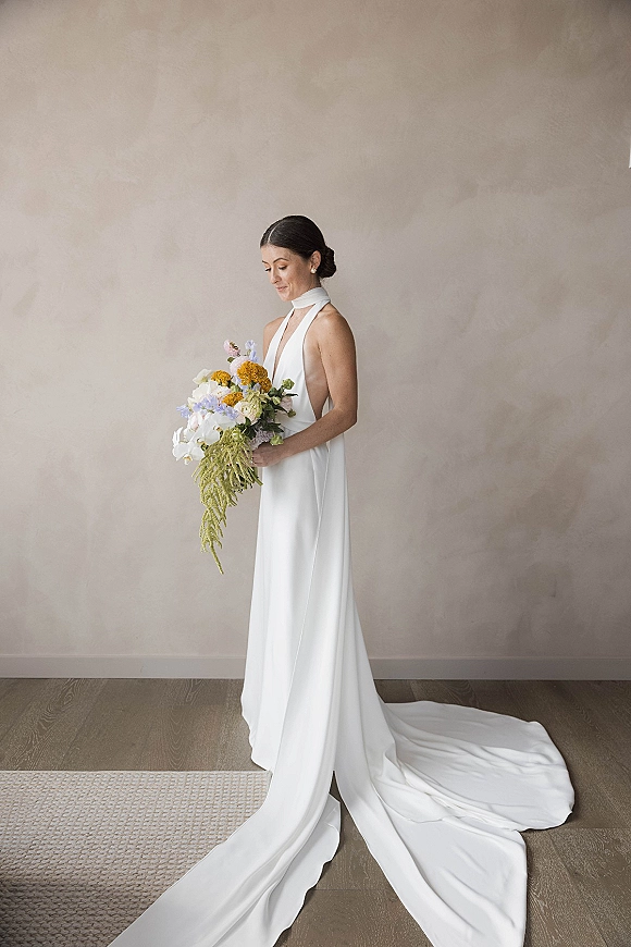 Bridal portrait of a bride in a halter neck wedding dress with a long train, holding an orchid bouquet with yellow flowers on a beige wall backdrop