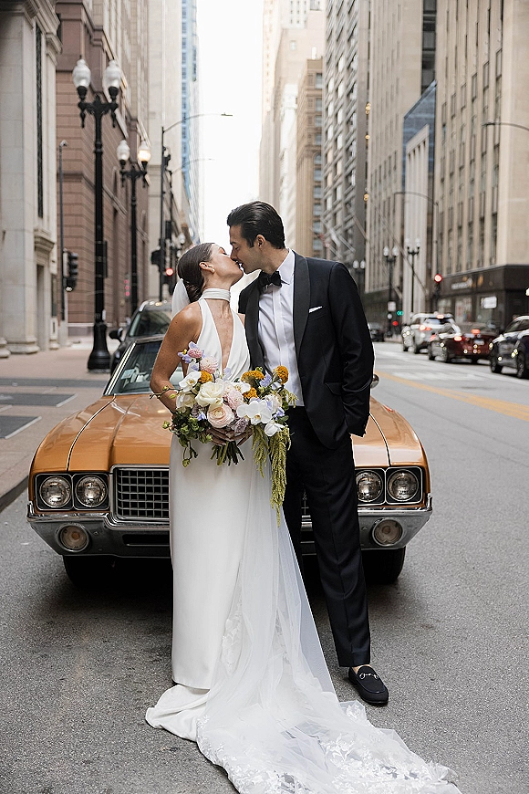 Wedding kiss portrait of bride and groom kissing beside a vintage car, her long veil and bouquet flowing on a downtown city street
