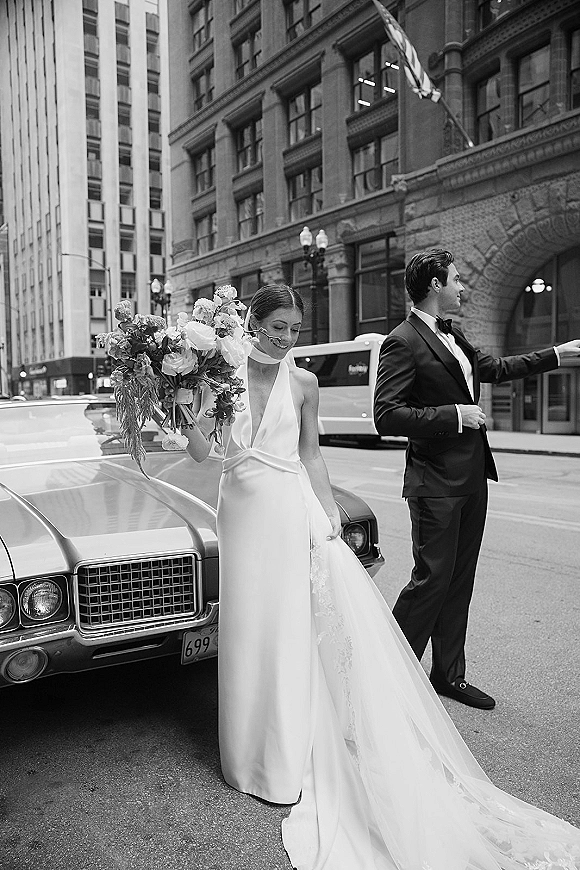 Couple portrait in a black and white wedding portrait style, bride with bouquet and lace train beside groom in tux by a vintage car on city street