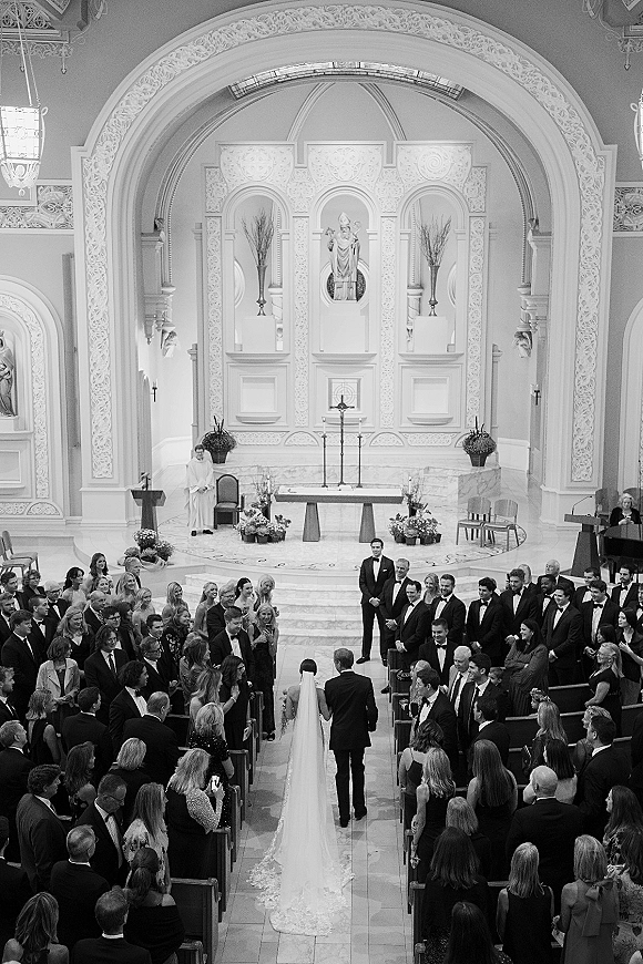 Wedding processional as bride walking down aisle with father, long veil and cathedral train, tuxedos ahead at ornate church altar with stained glass