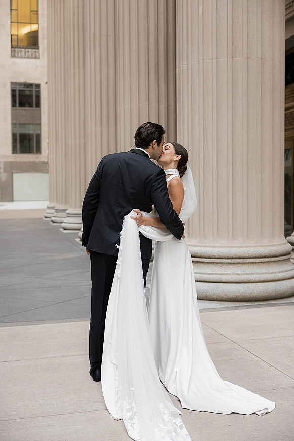 Wedding kiss portrait of the bride and groom kissing, her veil and gown train flowing as he holds it by stone columns downtown