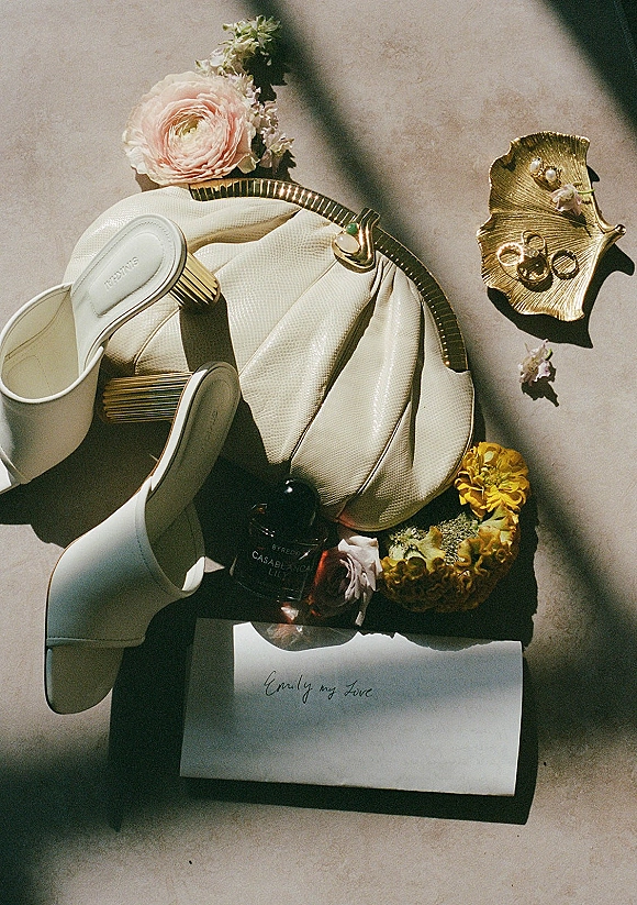 Wedding flat lay of white bridal shoes with perfume bottle and wedding rings on a stone surface, framed by flowers in sunlight shadows