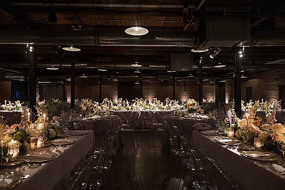 Reception tablescape with floral centerpieces and taper candles, place settings and glassware on a long table in an exposed brick industrial venue