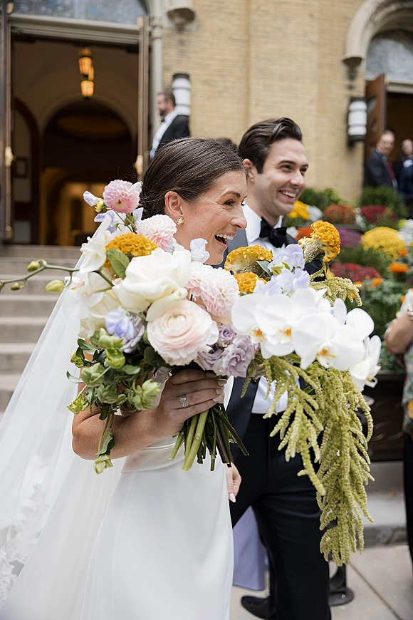 Wedding couple portrait with bride holding bouquet of white orchids and pastel blooms, smiling beside groom in tux on stone steps by brick entrance