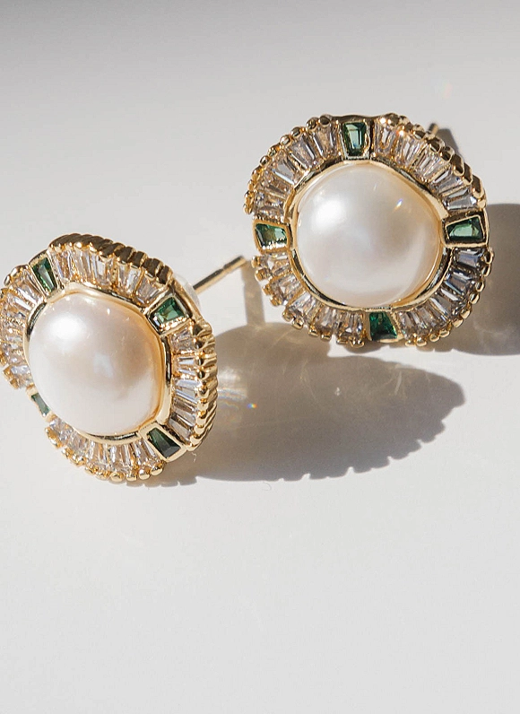 Bridal earrings, pearl stud earrings in gold settings with diamond halo and green gemstones, on a white surface with soft shadows