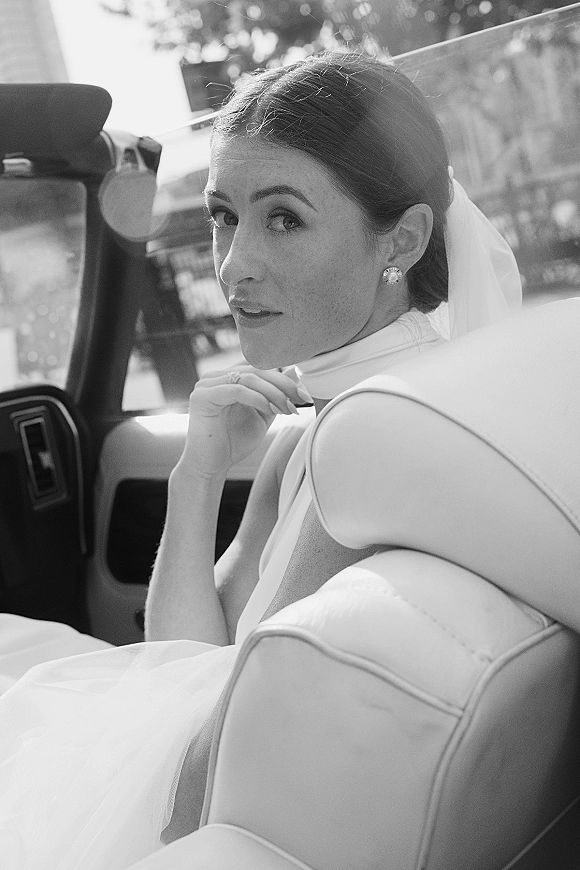 Bridal portrait of a bride in car looking over her shoulder, wearing a veil and pearl stud earrings by the window with greenery outside