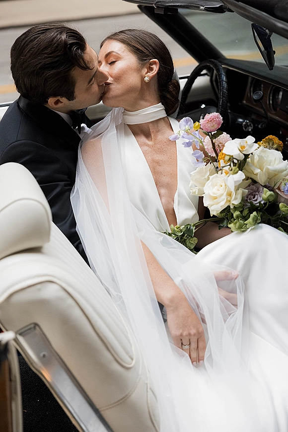 Wedding kiss portrait of bride and groom kissing in a convertible getaway car, bride in veil holding bouquet on a city street