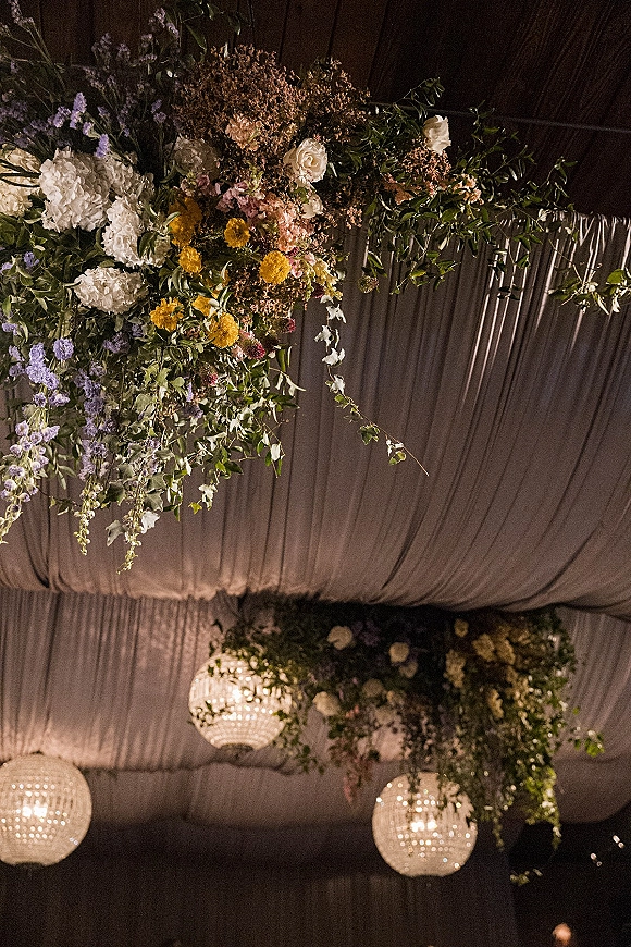 Reception ceiling decor with hanging floral installation of greenery and mixed blooms, draped fabric canopy and chandelier pendant lights in a dark tent