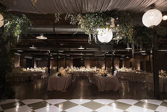 Reception decor in an industrial wedding reception with hanging greenery, chandeliers, candlelit long banquet tables, and a checkered dance floor