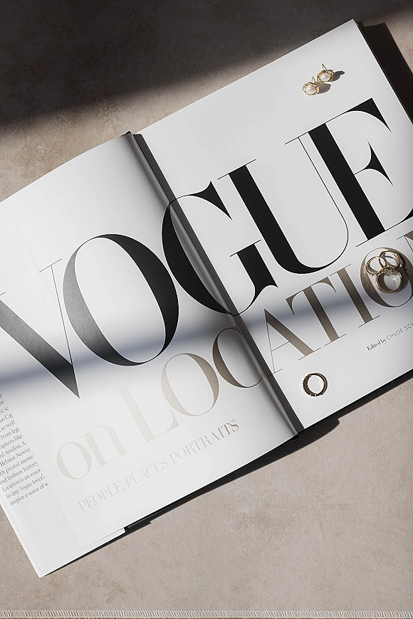 Wedding rings flatlay with gold rings beside pearl earrings on a magazine, styled on a stone surface with sunlit shadows