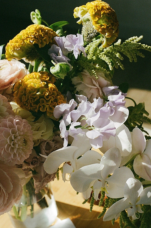 Wedding bouquet with white orchids in a glass vase, featuring yellow cockscomb, dahlias, and blush roses on a sunlit tabletop