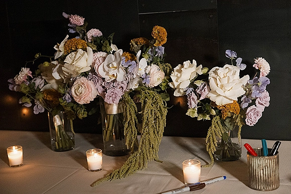 Wedding floral centerpieces and wedding bud vase centerpieces in glass vases with mixed blooms and tea lights on a tabletop against a dark wall