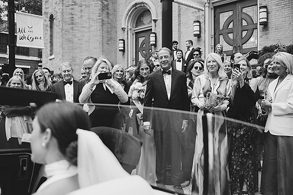 Wedding guest reaction as guests cheering and filming on phones in tuxedos and formal dresses outside a brick church doorway during exit