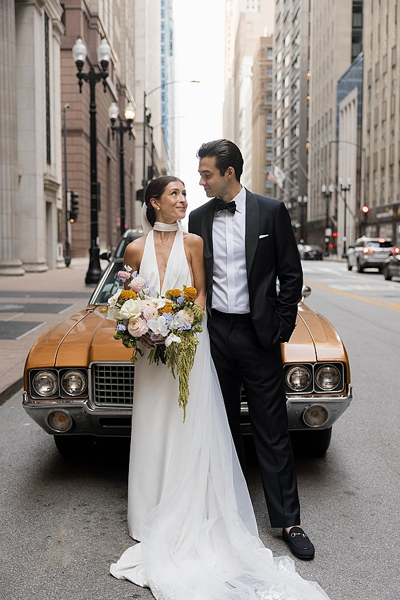Couple portrait of bride and groom leaning on a vintage car, bride holding a colorful bouquet, on a downtown city street with tall buildings