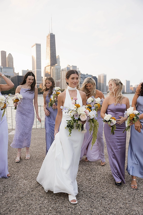 Bride with bridesmaids walking along the waterfront, holding colorful bouquets, city skyline behind them and a white halter gown in front