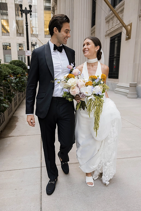 Couple portrait of bride and groom walking with a bouquet, her veil flowing beside a stone-columned city sidewalk backdrop