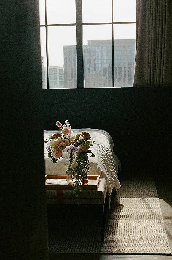 Wedding bouquet in a clear glass vase on a wooden tray atop bed linens, lit by window sunlight with city buildings beyond curtains