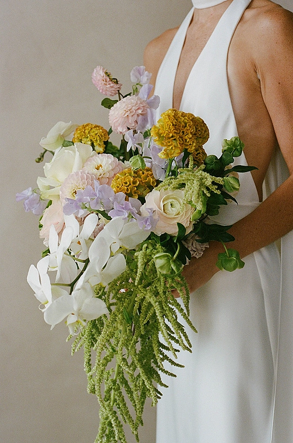 Bridal bouquet with cascading bridal bouquet greenery, orchids, roses, and pastel blooms tied with ribbon against a neutral wall