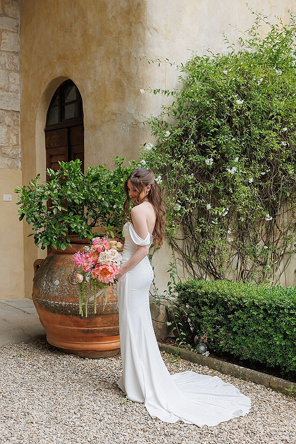Bridal portrait of a bride holding bouquet in side profile, wearing an off-the-shoulder gown with long train in a stucco courtyard.