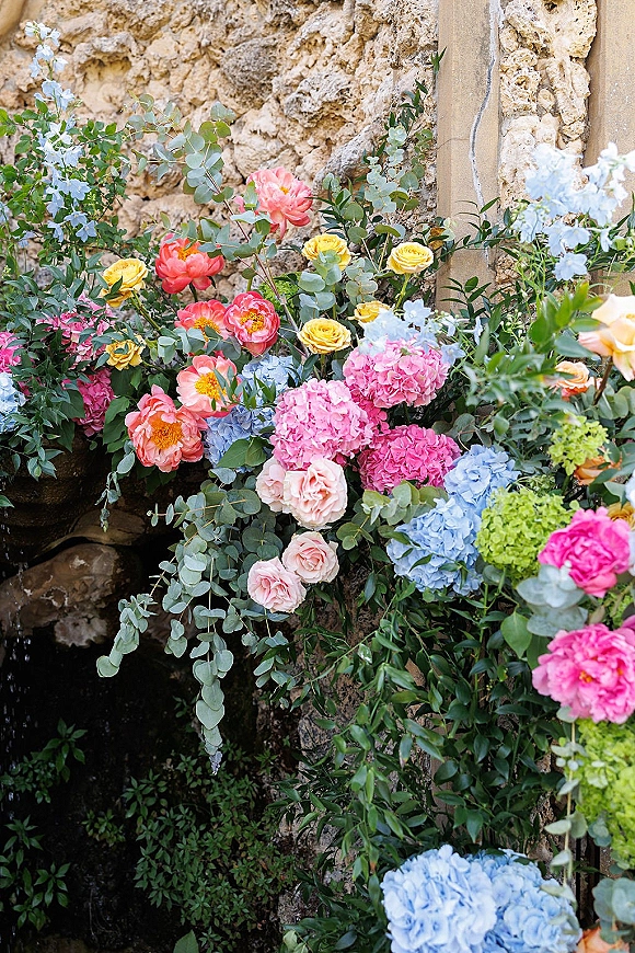 Wedding floral installation with a ceremony flower arrangement of roses, peonies, and hydrangeas spilling over a stone wall ledge