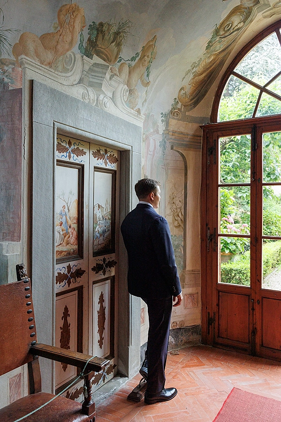 Groom portrait of a man in a navy suit looking out window by an arched window in a fresco-walled room with terracotta tile floor