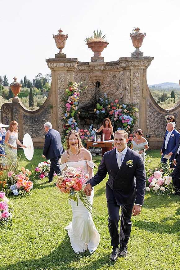 Wedding recessional as bride and groom walk the aisle hand in hand, she holds a colorful bouquet beside a stone fountain wall and guests
