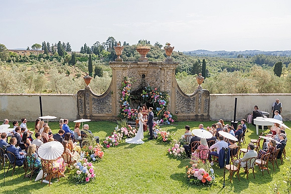 Outdoor wedding ceremony with a garden ceremony setup, featuring a floral arch, aisle flowers, white parasols, and grand piano by a stone fountain wall