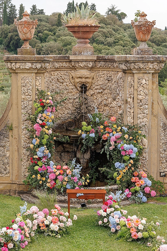 Ceremony backdrop with fountain wedding backdrop, roses and hydrangea garlands framing a vintage stone fountain against a stone wall lawn